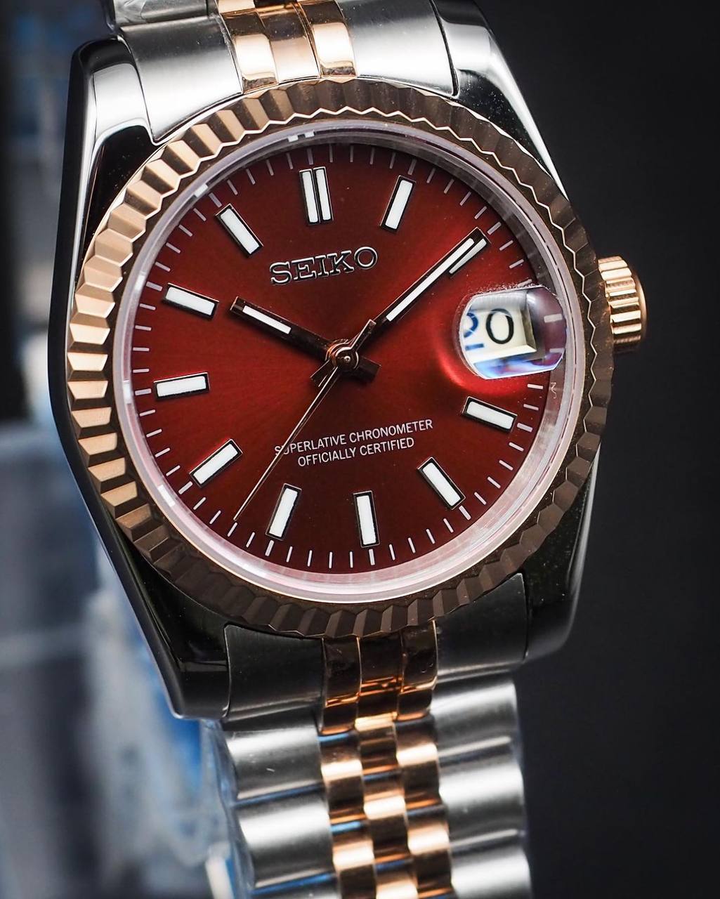 angled close-up of red sunburst dial Seiko mod watch showing date window, rose gold fluted bezel and two-tone bracelet